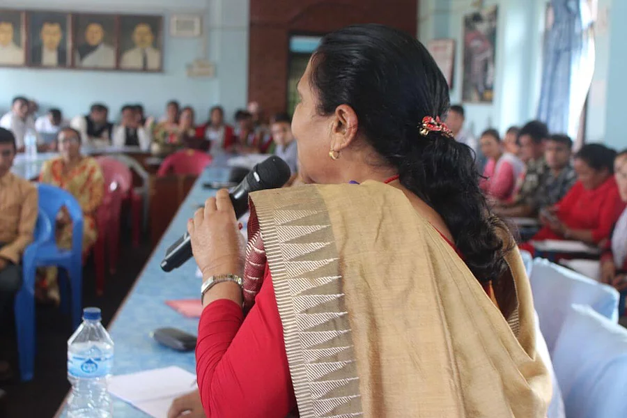 A young woman is speaking into a microphone in front of an audience at an event. She is wearing traditional clothing with a light-colored shawl draped over her shoulder.